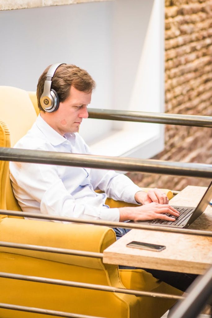 man-sitting-pn-yellow-chair-2451570 Adult man using laptop with headphones in a contemporary indoor workspace.